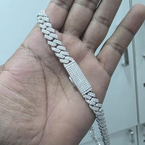 May include: A silver-colored cuban link chain bracelet with a rectangular clasp. The bracelet is encrusted with small, clear gemstones. The bracelet is held in a person's hand.