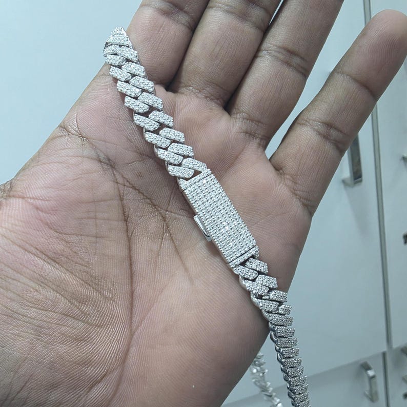 May include: A silver-colored cuban link chain bracelet with a rectangular clasp. The bracelet is encrusted with small, clear gemstones. The bracelet is held in a person's hand.