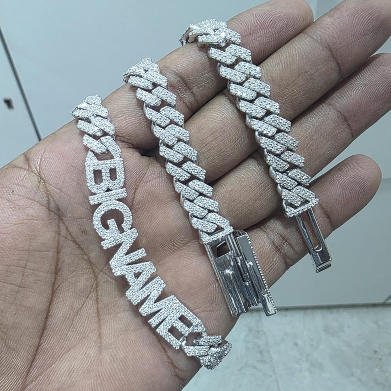 May include: Silver-toned bracelet with the word "BIGNAME" spelled out in large, sparkling, clear stones. The bracelet features a thick, link chain design, and a secure clasp. The jewelry is held in a hand.