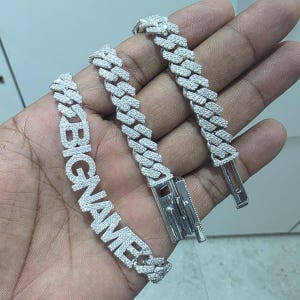 May include: A silver-colored bracelet with the word "BIGNAME" spelled out in large, sparkling letters. The bracelet is made of interlocking links, each encrusted with small, clear stones. The clasp is visible, and the bracelet is held in a hand.