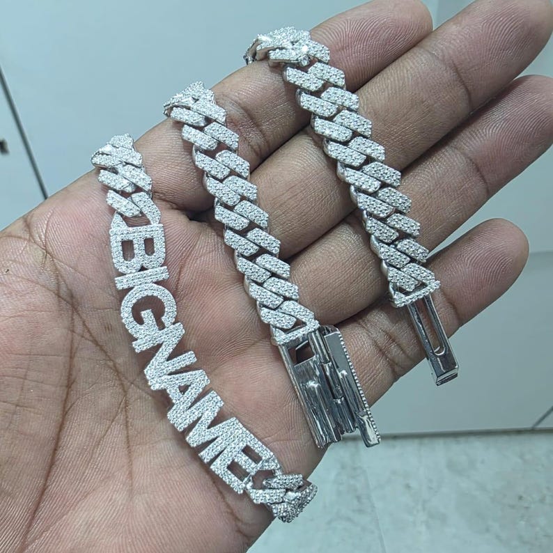 May include: A silver-colored bracelet with the word "BIGNAME" spelled out in large, sparkling letters. The bracelet is made of interlocking links, each encrusted with small, clear stones. The clasp is visible, and the bracelet is held in a hand.