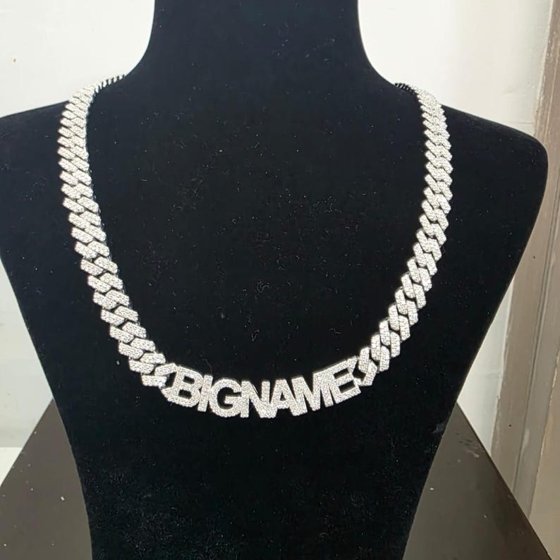 May include: A silver-colored cuban link chain necklace with the word "BIGNAME" spelled out in the center. The necklace is displayed on a black mannequin. The chain links are encrusted with small, sparkling stones.