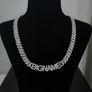 May include: A silver-colored chain necklace with the word "BIGNAMES" spelled out in large, block letters. The necklace is displayed on a black mannequin. The chain links are covered in small, sparkling stones.