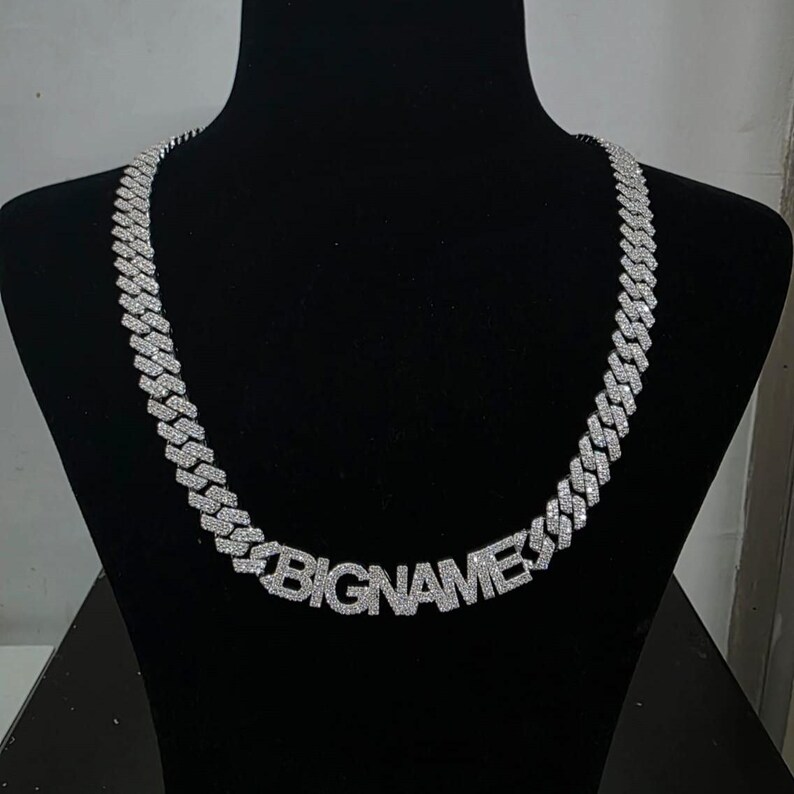 May include: A silver-colored chain necklace with the word "BIGNAMES" spelled out in large, block letters. The necklace is displayed on a black mannequin. The chain links are covered in small, sparkling stones.