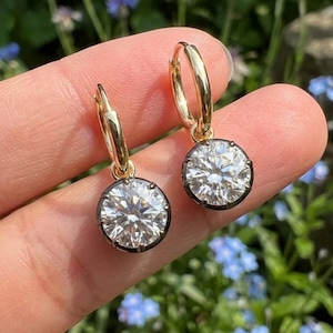 May include: A pair of gold hoop earrings featuring round, clear, faceted stones set in a dark metal bezel. Each earring consists of a polished gold hoop connected to a circular pendant that holds a brilliant-cut stone, reflecting light with clarity.