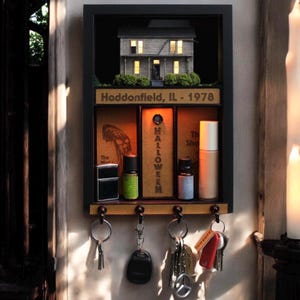 May include: A black framed key holder with a miniature house on top. The key holder has three vertical slots with the word "HALLOWEEN" in the center. The text "Haddonfield, IL - 1978" is above the slots. Key rings hang below.