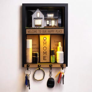 Puede incluir: Un organizador de pared con marco negro con una escena de casa en miniatura en la parte superior. El organizador tiene un estante con productos de belleza y un llavero con llaves y accesorios. El texto "WELCOME TO OUR HAPPY PLACE" está encima del estante.