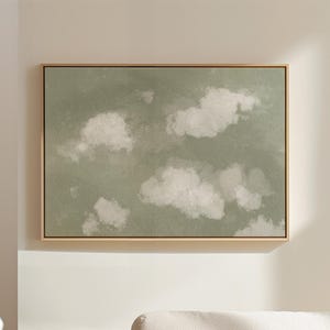 May include: A framed canvas art piece featuring a serene cloudscape. The artwork showcases fluffy white clouds against a muted green backdrop, encased in a light-colored wooden frame. The piece is mounted on a white wall, enhancing the artwork's visual appeal.