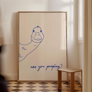 May include: A framed art print featuring a blue line drawing of a duck with the text "are you pooping?" The artwork is displayed in a room with a small wooden stool and a checkered floor. The frame is a light brown color.
