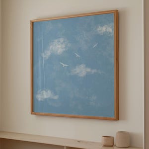 Op de afbeelding: Een ingelijst kunstwerk met een blauwe lucht met witte wolken en drie vogels in vlucht. Het kunstwerk is gevat in een lichtbruine houten lijst en hangt aan een crèmekleurige muur. Een sereen kunstwerk met een natuurthema.