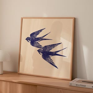 May include: A framed print featuring two stylized blue swallows in flight against a beige background. The artwork is displayed on a wooden cabinet, adding a touch of nature-inspired decor to the room. The print is in a wooden frame.