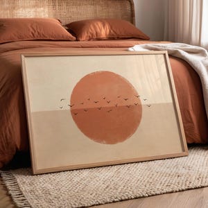 Op de afbeelding: Een ingelijste kunstprint met een grote oranje zon boven een horizon met vliegende vogels. Het kunstwerk staat op een bed met roestkleurig beddengoed, een rotan hoofdbord en een beige vloerkleed. De kamer heeft een warme, uitnodigende uitstraling.
