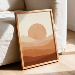 Peut inclure: Une impression d'art encadrée représentant un paysage stylisé avec des collines superposées dans des tons terre cuite, orange brûlé et crème. Un grand soleil circulaire uni de couleur orange sourd est centré au-dessus de l'horizon.