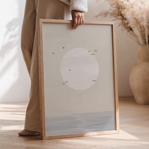 May include: A framed art print featuring a minimalist design with a large, pale circle resembling the sun, small black bird silhouettes, and a soft blue horizon. The frame is made of light-colored wood.
