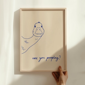 May include: A framed print featuring a whimsical blue line drawing of a duck. The duck is peeking from the side, with the text "are you pooping?" written below in a matching blue script. The frame is a light wood color.