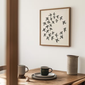 May include: A framed print of black birds in a circular formation hangs on a white wall. A wooden table below displays a grey mug, a dark green mug on a plate, and a ceramic vase. The room has a minimalist aesthetic.