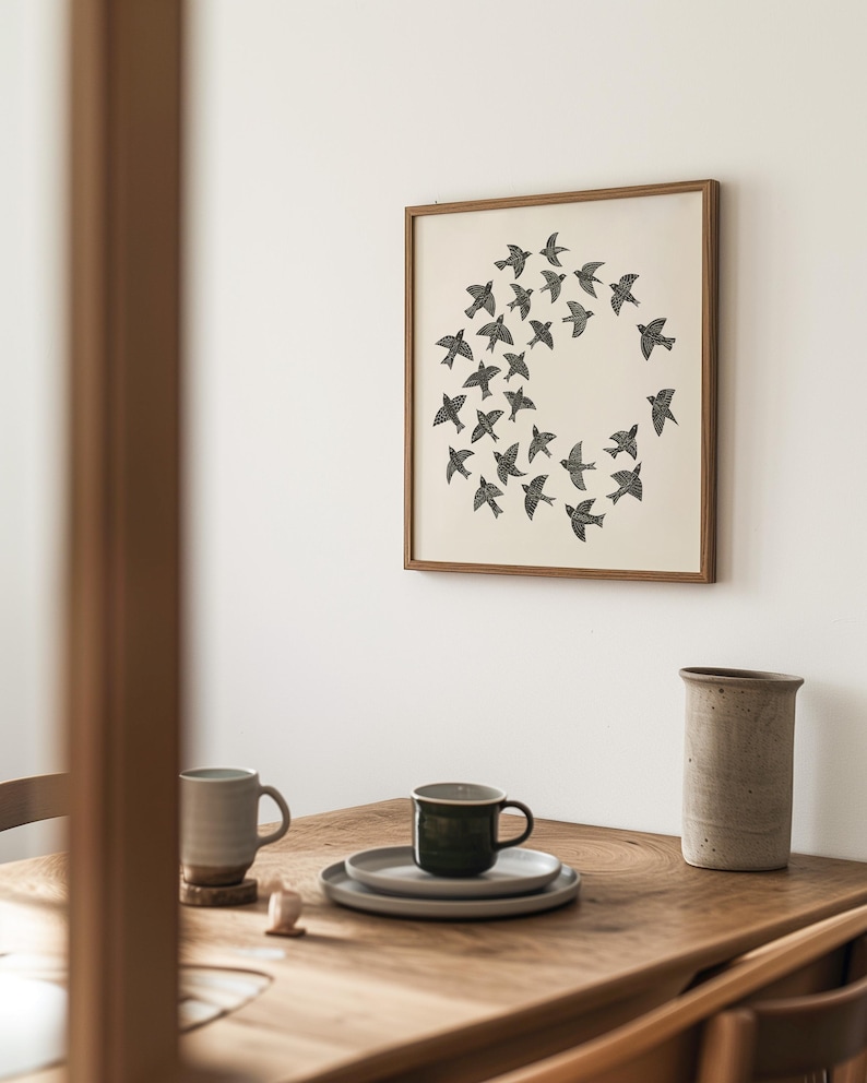 May include: A framed print of black birds in a circular formation hangs on a white wall. A wooden table below displays a grey mug, a dark green mug on a plate, and a ceramic vase. The room has a minimalist aesthetic.