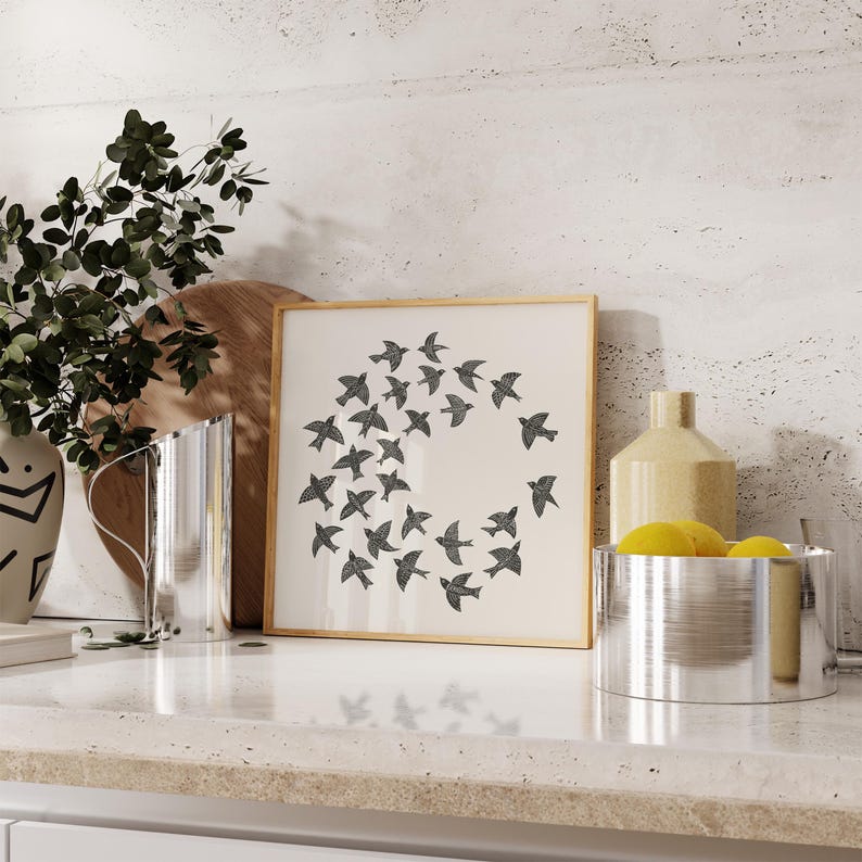 May include: A framed print with a design of black birds in a circle on a cream background. The artwork is on a white countertop, with a vase of greenery, a wooden board, and a metal container. The scene has a neutral colour palette.