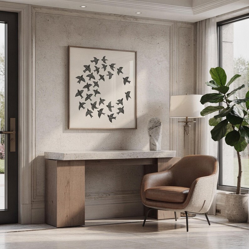 May include: A framed print showing a circular arrangement of black birds. The artwork is above a wooden console table with a marble top. A brown leather armchair and a potted plant are also in the room.