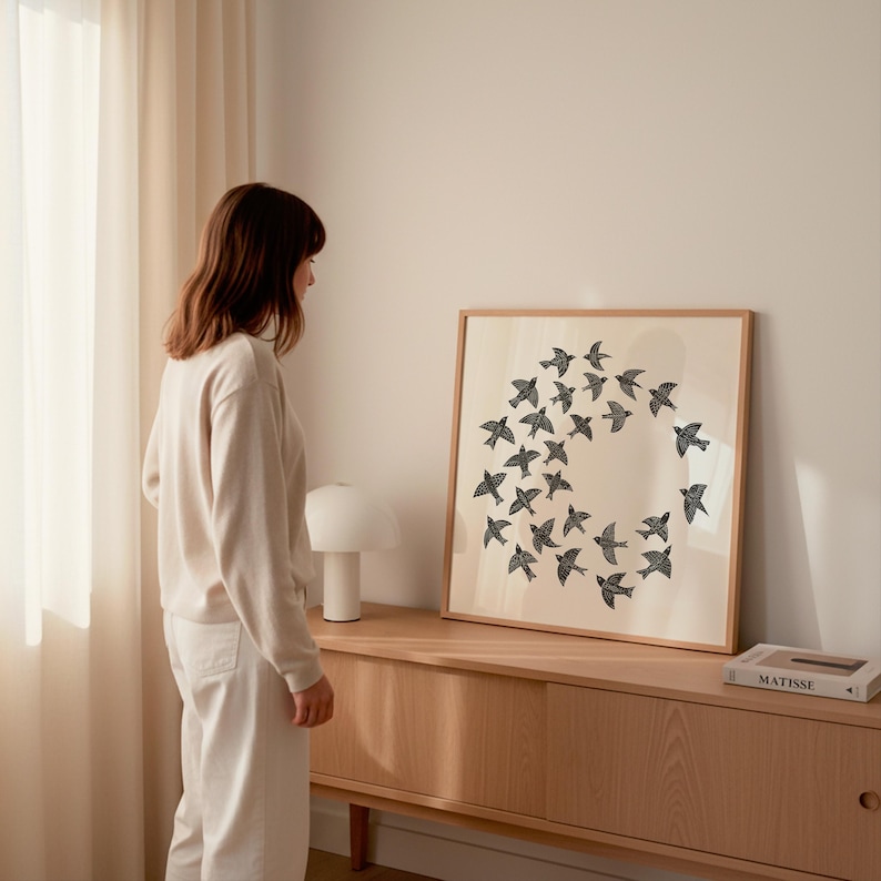 May include: A framed art print showing a circular arrangement of black birds on a cream background. The print is displayed on a wooden cabinet, with a lamp and a book. The room has a neutral colour palette with a person standing nearby.