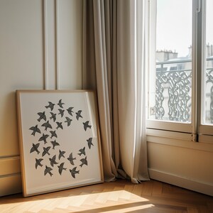 May include: A framed art print showing a circular arrangement of black birds on a white background. The frame is light wood. The print is propped against a wall near a window with beige curtains, in a room with wooden flooring.