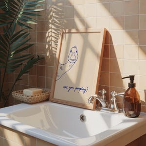 May include: A framed print with a cartoon duck illustration and the text "are you pooping?" in blue. The print is in a light wood frame, resting on a white sink. A brown soap dispenser and a woven basket are also visible.