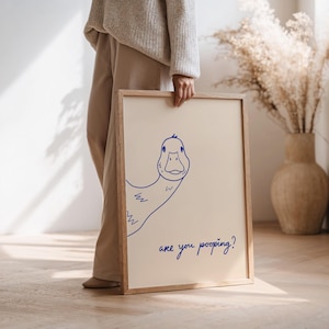 May include: A framed art print featuring a blue line drawing of a duck with the text "are you pooping?" on a cream background. The artwork is in a light wood frame.