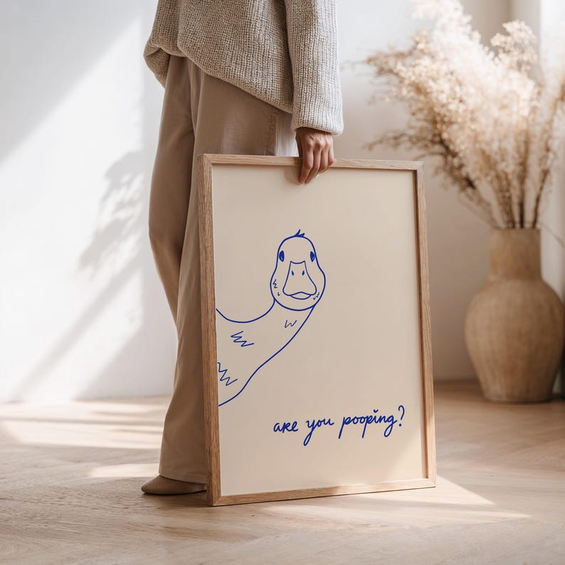 May include: A framed art print featuring a blue line drawing of a duck with the text "are you pooping?" on a cream background. The artwork is in a light wood frame.