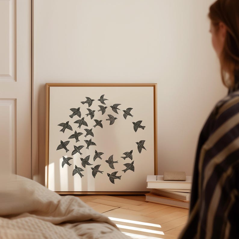 May include: A framed print showing a circular arrangement of black bird silhouettes on a cream background. The artwork is displayed in a room with a wooden floor and neutral-toned walls.