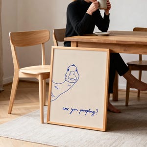 May include: A framed print with a cartoon duck illustration in blue ink. The duck is looking at the viewer. The text "are you pooping?" is written in blue ink below the duck. The frame is made of light-colored wood.