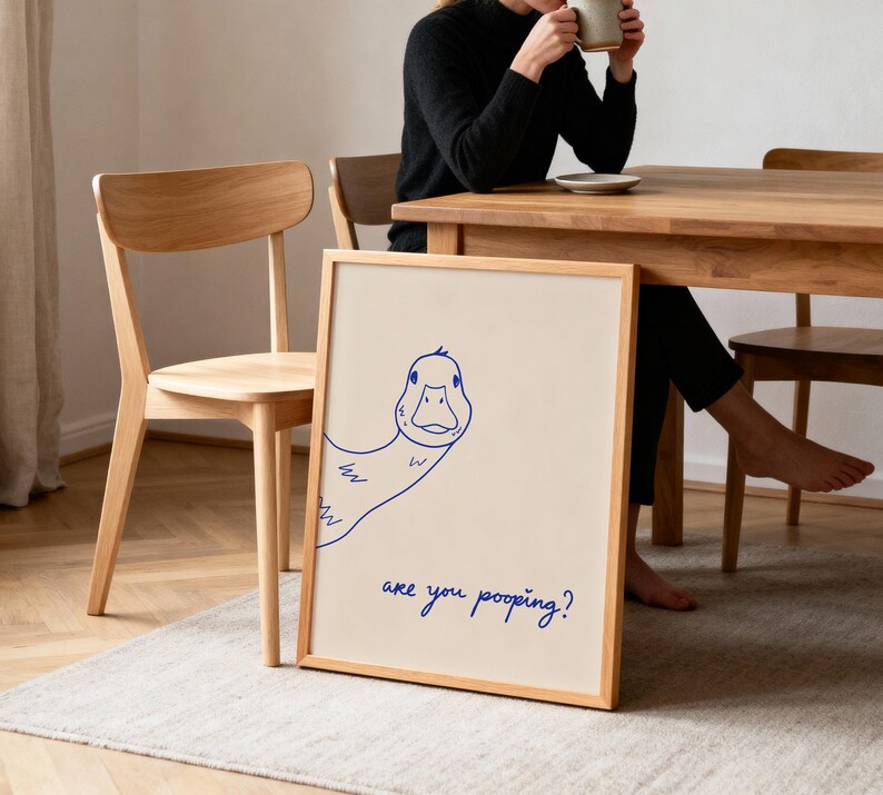 May include: A framed print with a cartoon duck illustration in blue ink. The duck is looking at the viewer. The text "are you pooping?" is written in blue ink below the duck. The frame is made of light-colored wood.