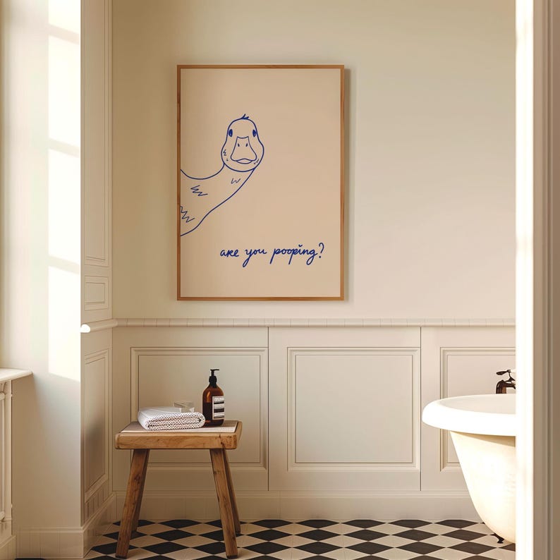 May include: A framed art print featuring a whimsical blue line drawing of a duck with the text "are you pooping?" The artwork is displayed in a bathroom setting, with a wooden stool, towels, and a bottle of soap.