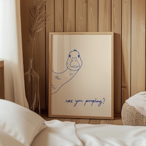 May include: A framed print featuring a blue line drawing of a duck peeking from the side. The text "are you pooping?" is written in blue script below the duck. The artwork is displayed in a light-colored wooden frame.
