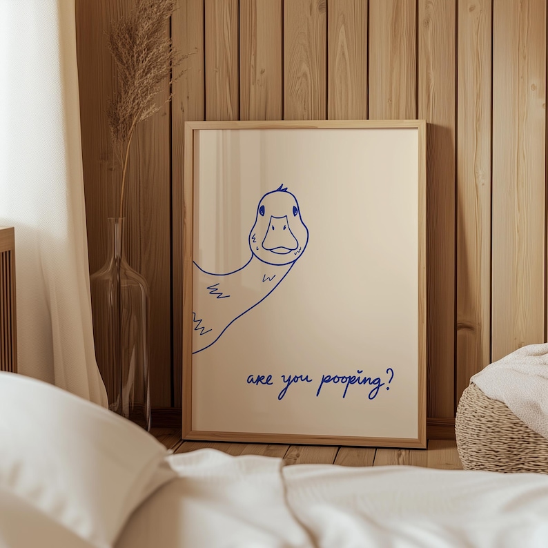 May include: A framed print featuring a blue line drawing of a duck peeking from the side. The text "are you pooping?" is written in blue script below the duck. The artwork is displayed in a light-colored wooden frame.