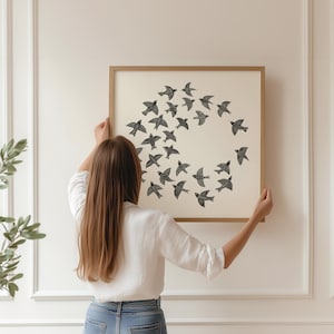 May include: A framed print showing a circular arrangement of black birds on a cream background. The artwork is held up against a white wall. The frame is a light brown colour. The image suggests a theme of nature and home decor.