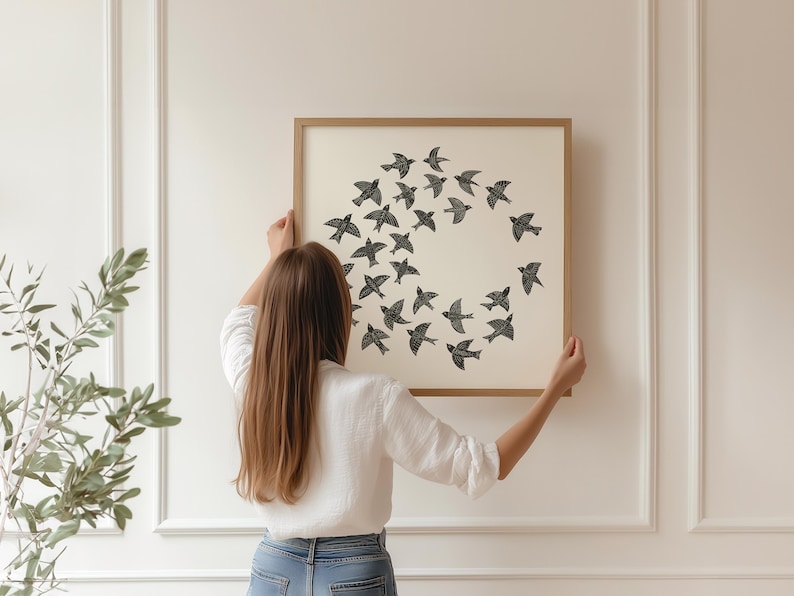 May include: A framed print showing a circular arrangement of black birds on a cream background. The artwork is held up against a white wall. The frame is a light brown colour. The image suggests a theme of nature and home decor.