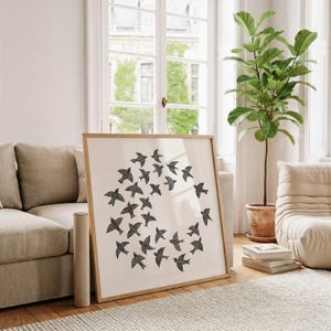 May include: A framed print with a design of black birds in a circle on a cream background. The artwork is in a light wood frame, placed against a beige sofa in a bright room. A rolled canvas and a potted plant are also visible in the room.