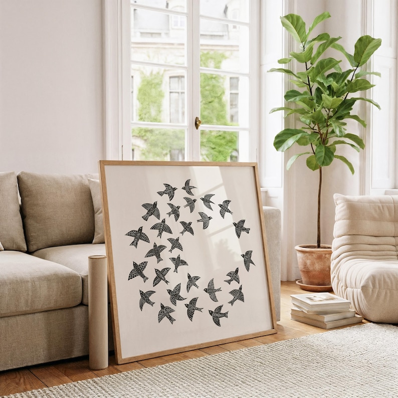 May include: A framed print with a design of black birds in a circle on a cream background. The artwork is in a light wood frame, placed against a beige sofa in a bright room. A rolled canvas and a potted plant are also visible in the room.