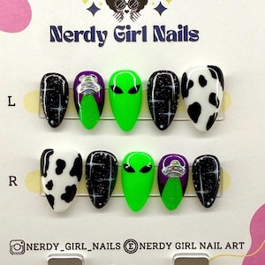 May include: A set of Nerdy Girl Nails press-on nails. The nails feature various designs, including black glitter, a green alien face, a purple alien spaceship, and a black and white cow print. The brand name and social media handles are visible.