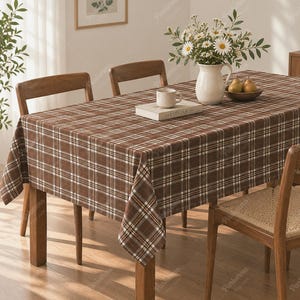 May include: A rectangular tablecloth featuring a brown, white, and tan plaid pattern draped over a wooden dining table. The table is surrounded by wooden chairs with woven seats and holds a white ceramic pitcher with daisies and a bowl of pears.