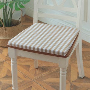 May include: A white wooden chair featuring a square seat cushion with a beige and white gingham check pattern. The chair has a slatted backrest design and is positioned on a wooden floor next to a potted plant in a woven basket container.