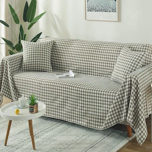 May include: A sofa draped with a brown and white gingham patterned throw. Two matching cushions are on the sofa. A small white table with a plant and a lemon sits in front.