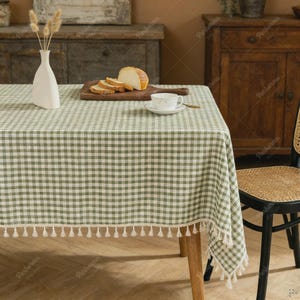 Puede incluir: Un mantel a cuadros verdes y blancos con un ribete de borlas blancas. Un jarrón blanco con flores secas, una tabla de cortar de madera con pan en rodajas y una taza y platillo están sobre la mesa. La mesa tiene patas de madera.