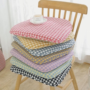 Puede incluir: Una pila de siete cojines cuadrados para silla con esquinas redondeadas, cada uno con un patrón de cuadros vichy de diferente color en rosa, amarillo, azul, morado, azul claro y negro, coronado por una taza de cerámica blanca.