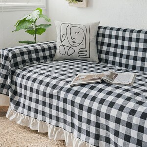 Könnte beinhalten: Eine schwarz-weiß karierte Sofahusse mit cremefarbenem Rüschenbesatz. Ein dekoratives Kissen mit einer Strichzeichnung eines Gesichts liegt auf dem Sofa. Eine offene Zeitschrift liegt auf dem Sofa. Eine grüne Pflanze befindet sich im Hintergrund.