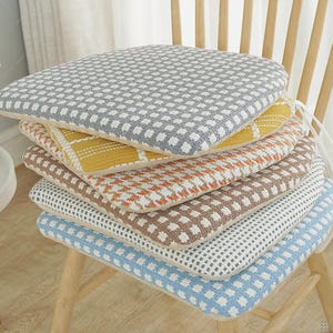 May include: A stack of five square chair cushions with rounded corners, each featuring a distinct woven grid pattern in colours including grey, yellow, orange, brown, and blue, all with white accents and white fabric ties attached to the sides.