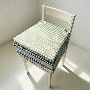 Peut inclure: Une pile de quatre coussins d'assise carrés avec un motif pied-de-poule dans des tons vert, noir, bleu marine et marron. Chaque coussin présente un design épais et rembourré avec des coins arrondis, posé sur une chaise en bois clair.