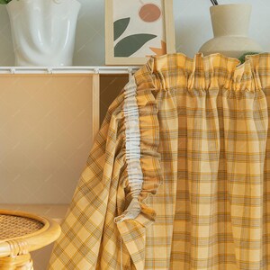 May include: Yellow plaid curtain with a white ruffled edge. The curtain is hanging in front of a light-colored cabinet. A framed picture and a white vase are visible in the background.