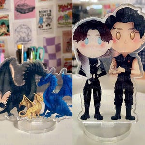 Puede incluir: Expositores acrílicos con ilustraciones de dibujos animados. Uno muestra un par de personajes con trajes negros, el otro una familia de dragones en azul, negro y dorado. Los expositores tienen una base transparente.