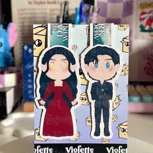 May include: A bookmark featuring two cartoon figures, a woman in a red dress and a man in a black suit. The bookmark has a light blue background with a pattern of small cheese slices. The text "Violette Signet" is visible.